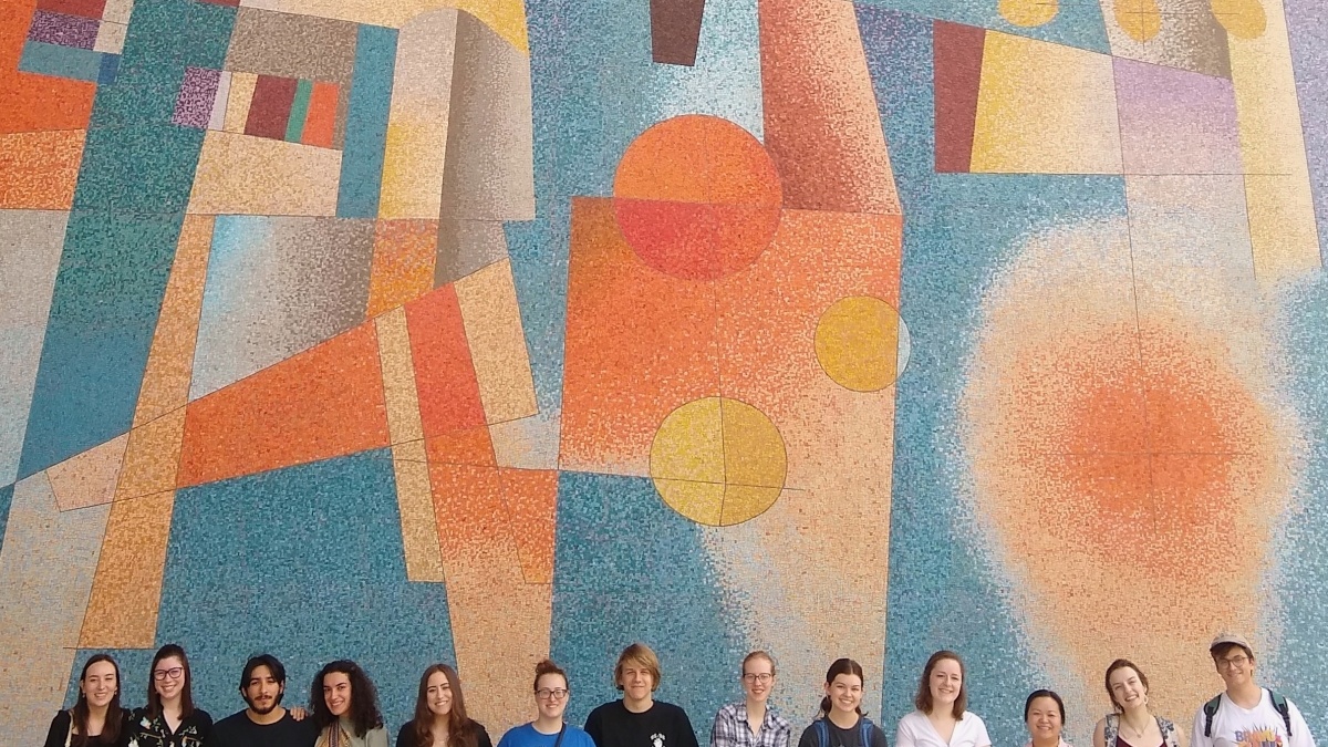 students stand collectively in front of a large glass mosaic by Carlos Merida