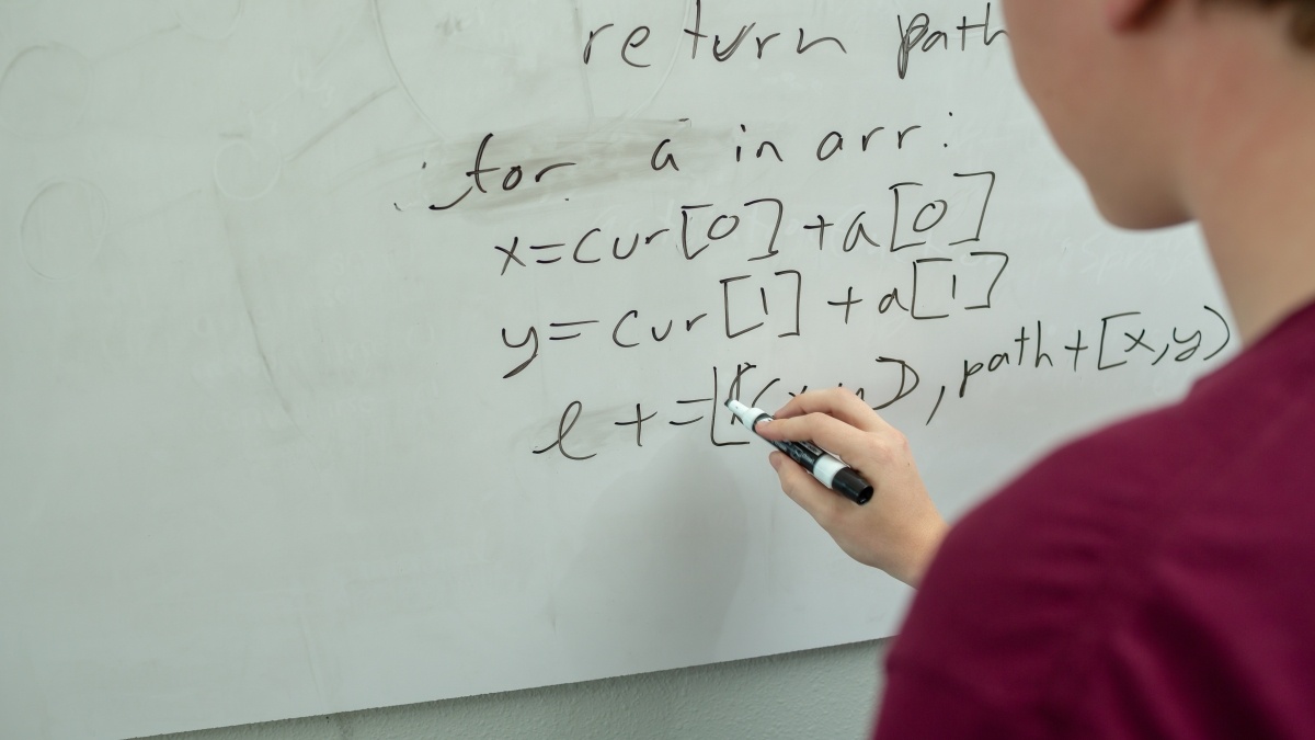 A student writes code and math on a whiteboard. 